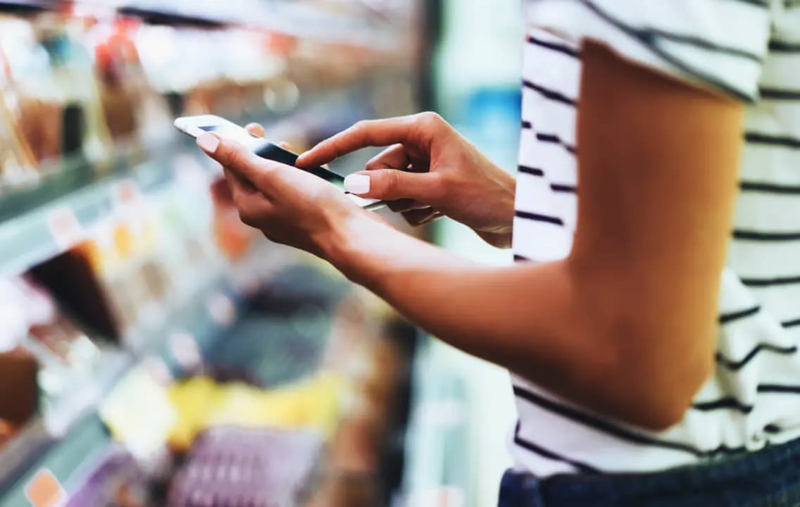 A woman checks her phone in a grocery store, utilizing in-store planning strategies to enhance margins and revenue. A woman checks her phone in a grocery store, utilizing in-store planning strategies to enhance margins and revenue.